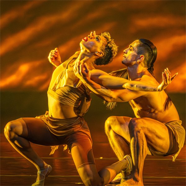 Two people kneel while dancing dramatically. The person in front is looking upward while opening their arms.
