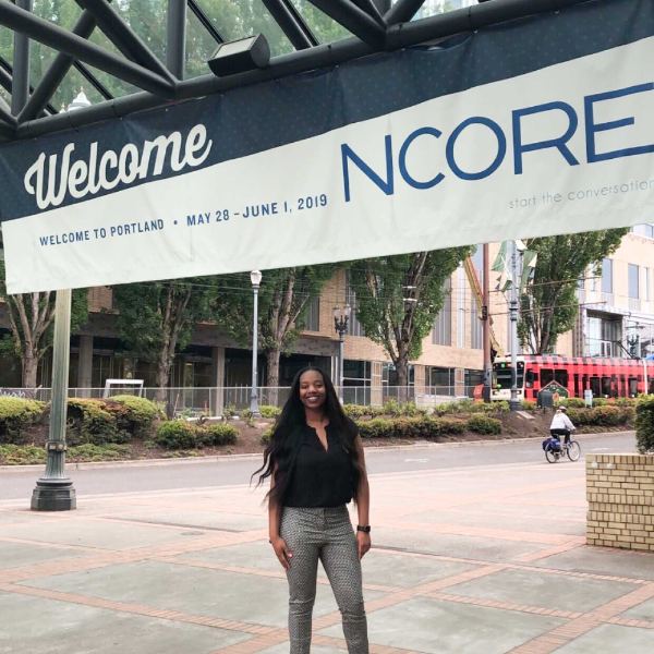 Jasmine Wilson is standing outside a conference in Portland Oregon under a banner that reads Welcome to NCORE.