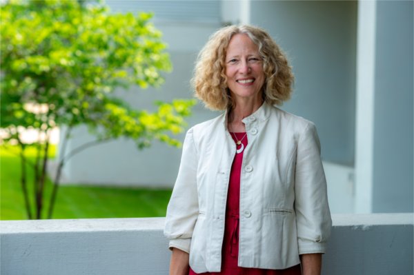 A person standing next to a short wall, with a tree in the background, smiles.