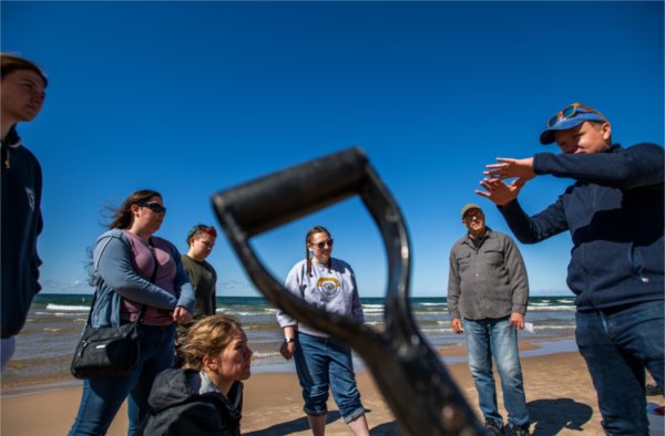 One person talks while others listen while standing on a Lake Michigan beach. The lake is in the background. One of the people is framed by a shovel handle in the foreground.