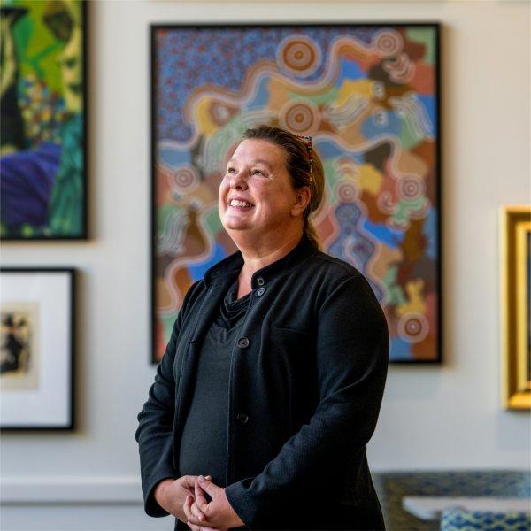 Melanie Shell-Weiss stands in front of a gallery wall of colorful paintings.