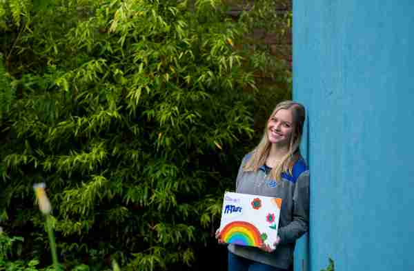 A person smiles while learning against a wall holding a sign with a rainbow and flowers.
