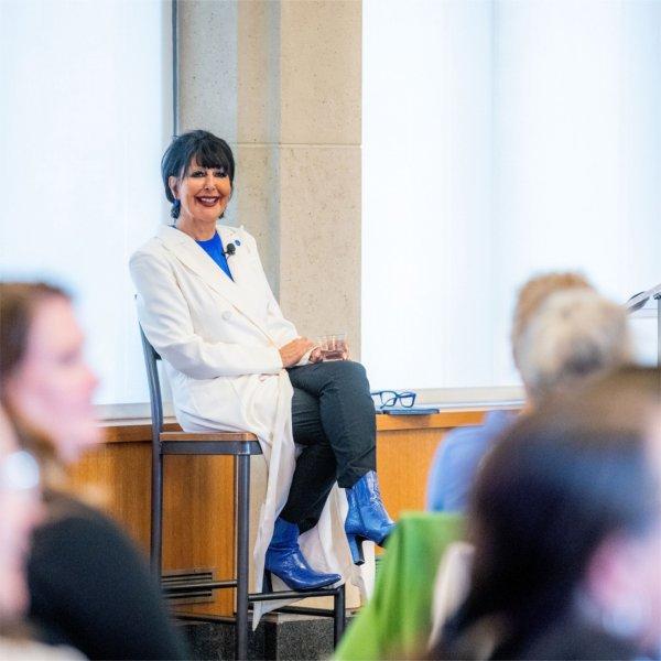 In a long white coat and blue heeled boots, President Mantella smiles as she sits at the front of a room and answers questions.