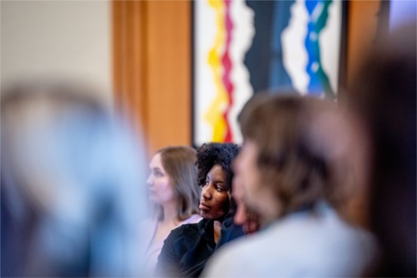 A woman sits at a table with others and watches President Mantella give a presentation. 