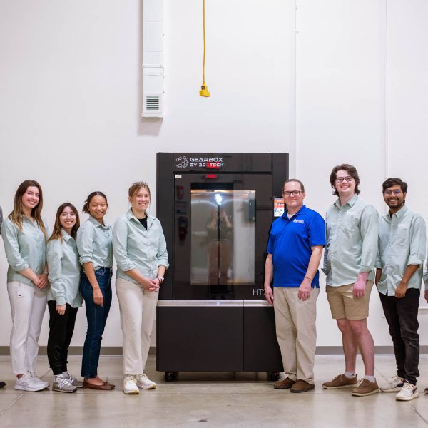 seven people, most in light green button down shirts, stand by a GearBox 3D printer in an industrial room in the Shape Corp. innovation design center