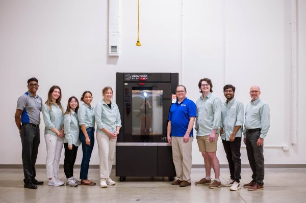 nine people, most in light green button down shirts, stand by a large 3D printer in the Shape Corp. Industrial Design Center