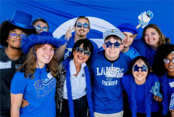  A university president wearing sunglasses smiles in the middle of a group of students all wearing blue. 