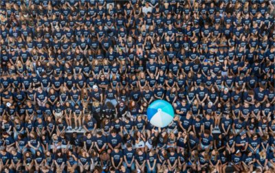 As seen from a drone, a large blue and white beach ball is tossed around by a stadium full of students. 