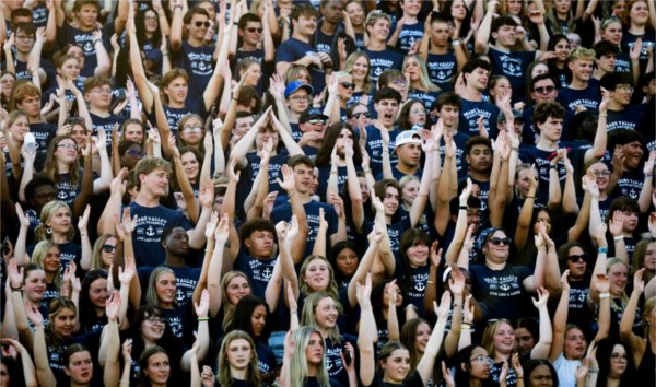  A stadium full of incoming college freshmen wearing matching blue shirts raise their hands in the air. 