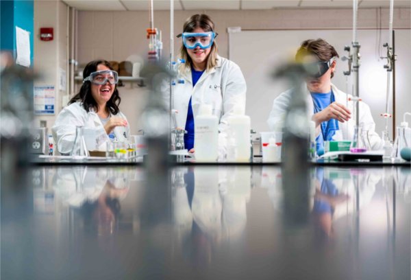  Three college students wearing white lab coats and goggles work in a chemistry lab setting. 