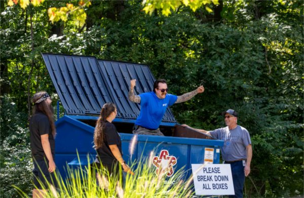  A person with a blue shirt and tattoos jumps up and down in a dumpster to smash down boxes. 
