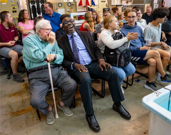Two men laugh together during an event as they sit among college students.  