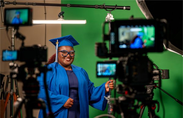 A portrait of a film and video college student standing with equipment in front of a green screen wearing a blue cap and gown smiles at the camera. 