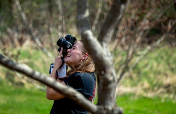  A college student with a camera to her face smiles as she takes photos of a tree. 