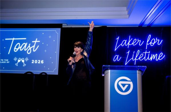  A university president tosses her hand into the air in front of a screen that reads Toast while a "Laker for a Lifetime" signs hangs nearby. 