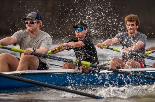 A row team practices on a river with water splashing in front of the boat. 