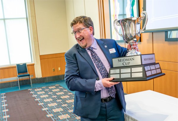 A person wearing a suit and tie smiles as they hold up a large Seidman Cup trophy. 