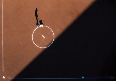 A birds eye view shows a softball field's pitcher's circle with a shadow of a person laying chalk lines on the dirt