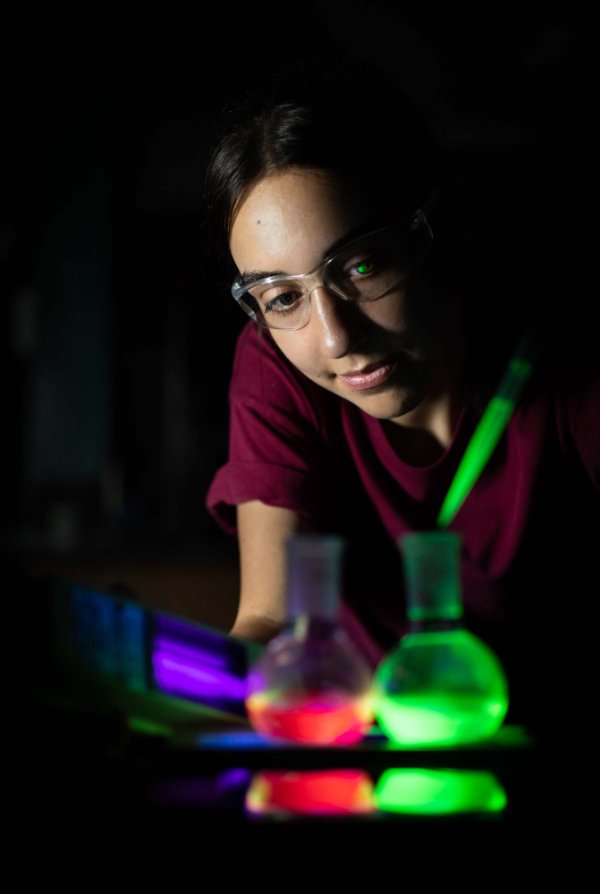 A person holding a light leans toward two beakers that glow in the foreground