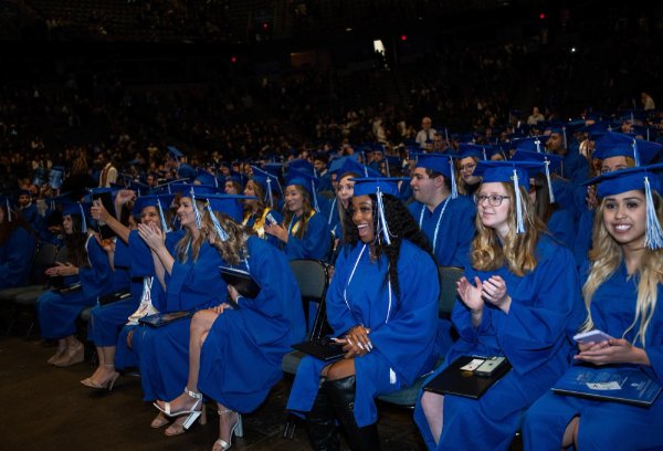 People in graduation garb smile while seated in an arena.