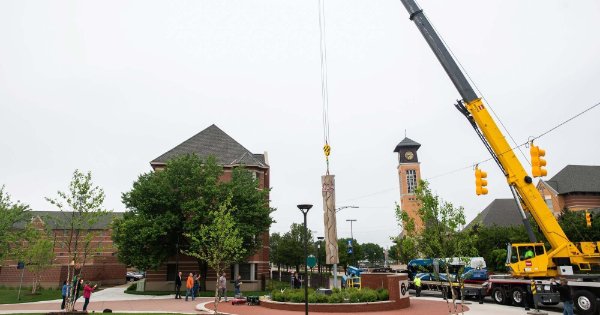 A vertical sculpture hanging from the cable of a crane is placed into its position.