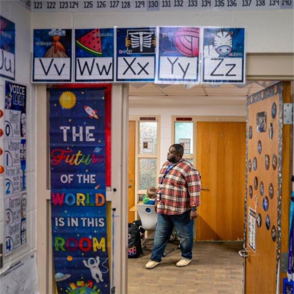 Charles Ball stands in hallway wearing a plaid shirt, his classroom door is adorned with letters of the alphabet and colorful space posters