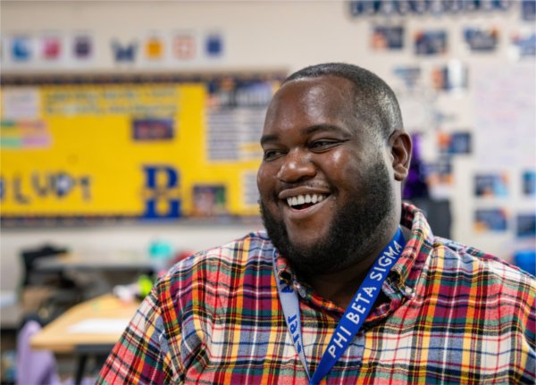 Charles Ball smiles in his classroom, wearing a plaid shirt and laynard