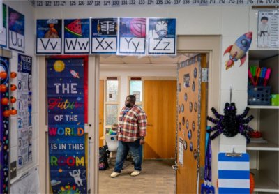 Charles Ball stands in hallway wearing a plaid shirt, his classroom door is adorned with letters of the alphabet and colorful space posters
