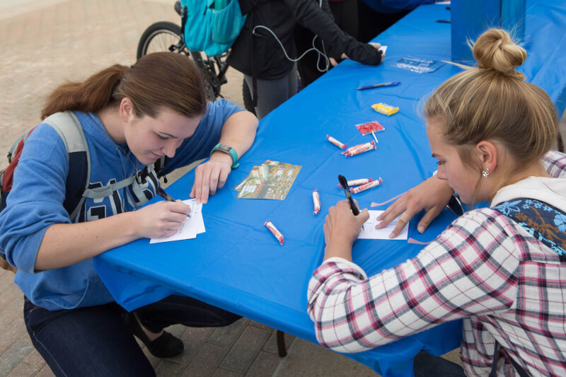  Female student wearing a plaid shirt kneeling while writing on a postcard