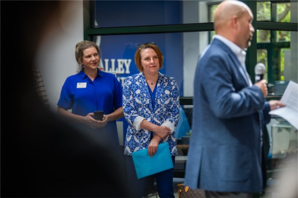 two women stand against a wall, man with microphone at right