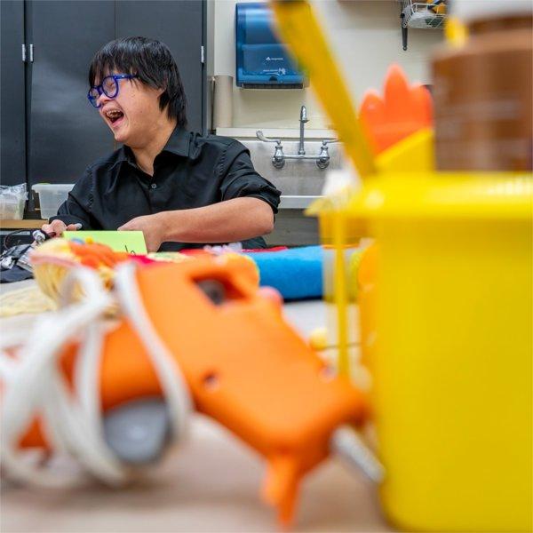 An individual laughs, with a hot glue gun and craft bin seen in the foreground.