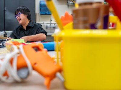 An individual laughs, with a hot glue gun and craft bin seen in the foreground.