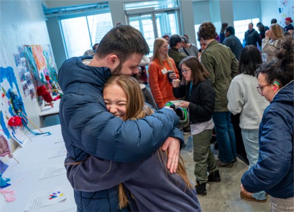 Two individuals hug joyfully while standing in an art gallery filled with others.