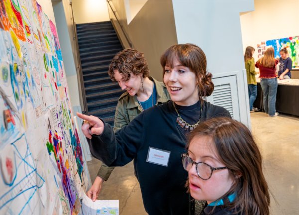 Three individuals look at the collage of drawings on the wall, with one pointing to a drawing.