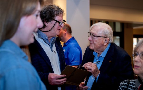 Don Lubbers at right signing an autograph for a man with glasses on a chain around his neck, two women standing close by