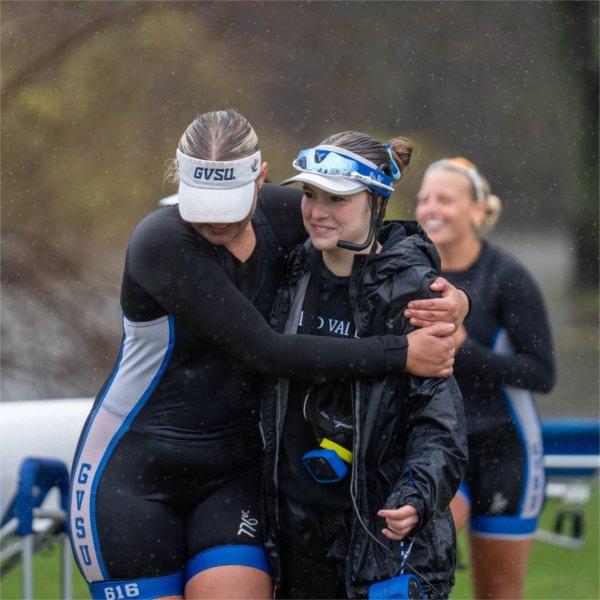two rowers hug after a race, they are in black uniforms, person on right wears a headset as the coxswain