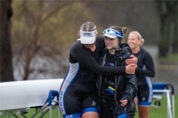 two rowers hug after a race, they are wearing black uniforms, person on right wears a headset as the coxswain