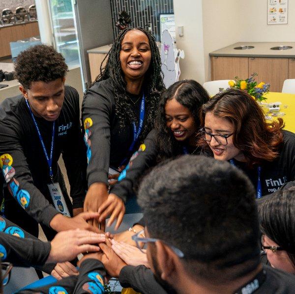 A group of students wearing matching black Fin Lit shirts stand in a circle and hold their hands together in the center.