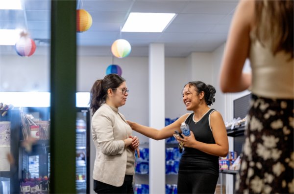 Two women stand in Replenish and chat. One places her hand on the other&#8217;s shoulder. 