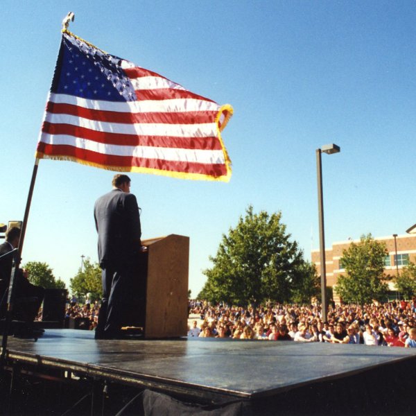 A person stands on a stage while speaking at a podium to a large crowd. An American flag flies behind the person.