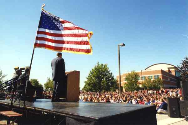 Person on a stage speaks at a podium to a large crowd. An American flag is flying behind the person.