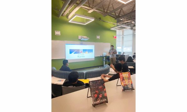 A Van Andel Institute staff member leads a presentation in front of a room of high school students. Two library books are in the foreground.