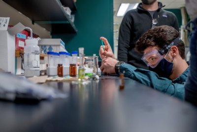 A student wearing goggles and a mask squats to closely measure a substance in a laboratory.