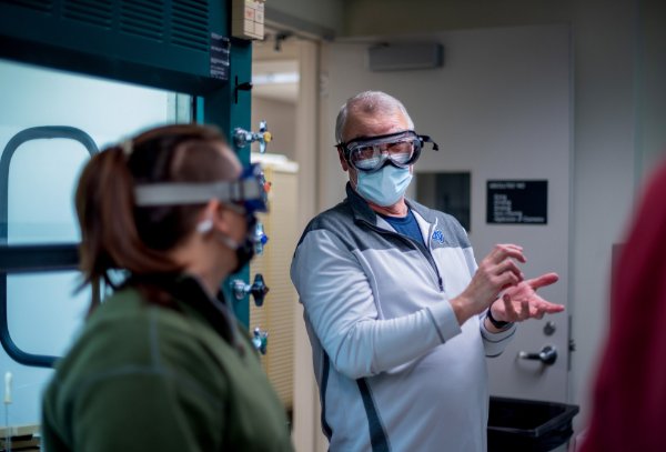 A faculty member gestures while talking to another person. Both are wearing goggles.
