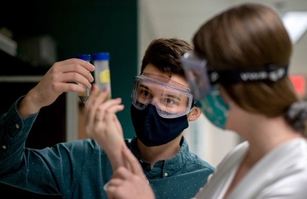 Two students, both wearing goggles and masks, study the contents of a test tube.