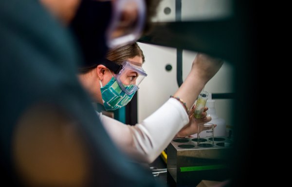 A student wearing goggles and a mask holds a container while working with a substance in the chemistry lab.