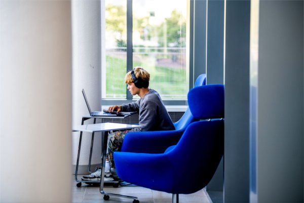 A college student works on their laptop while wearing headphones as they sit in a large blue chair. 