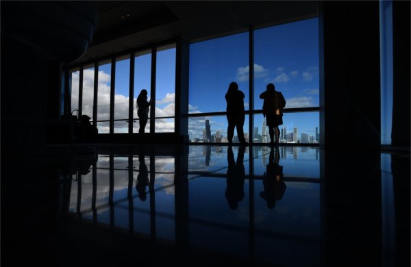Several people silhouetted against large glass windows with a city skyline behind them. 