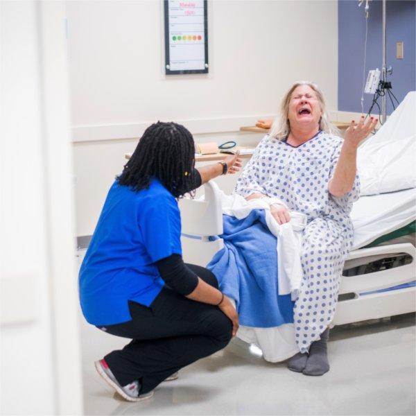 nursing student in blue scrub top kneels before a standardized patient who is in a hospital bed