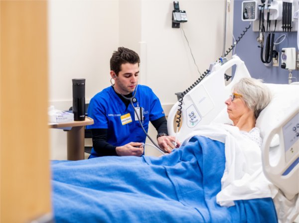 nursing student in blue scrub top takes the blood pressure reading of a patient in a hospital bed